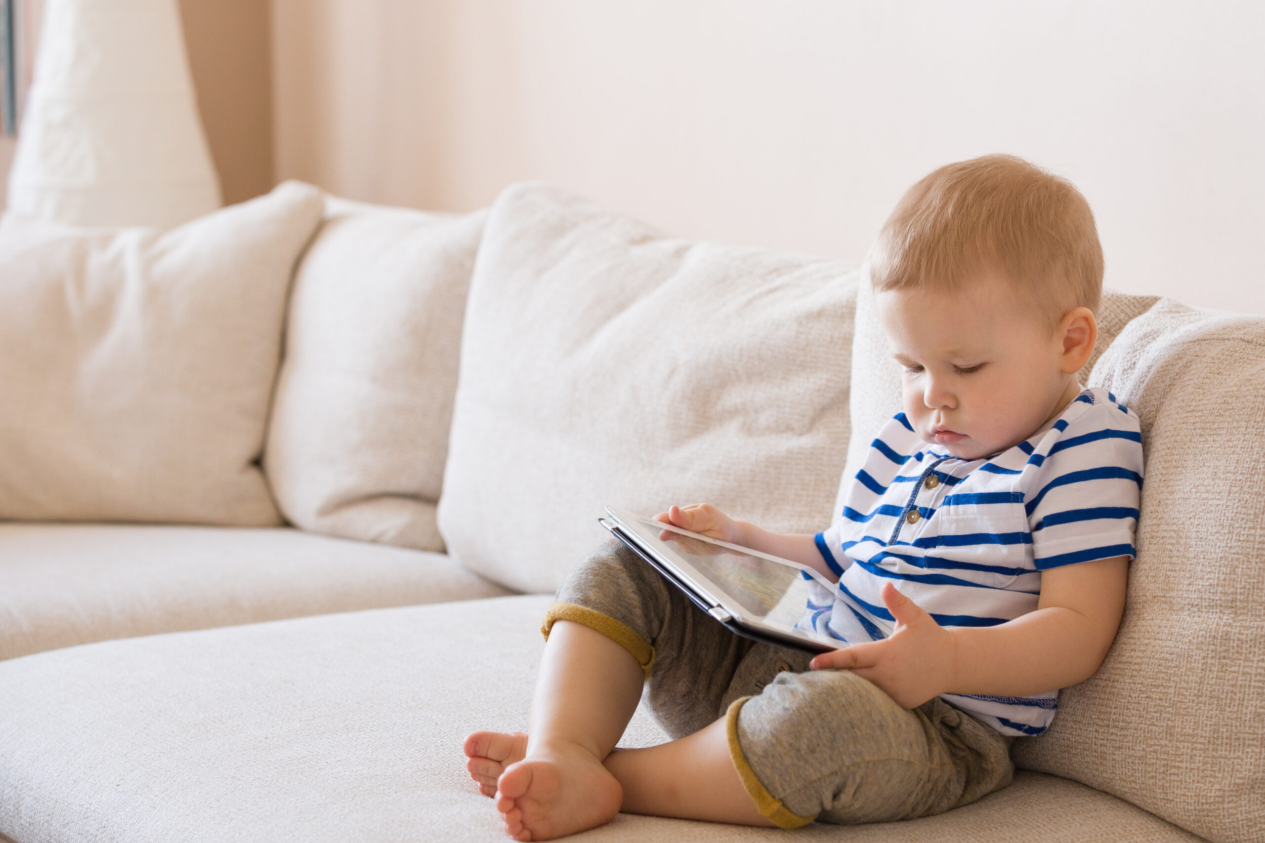 Adorable blond toddler boy laying on the sofa and playing with tablet pc at home, indoors. Child with tablet computer.